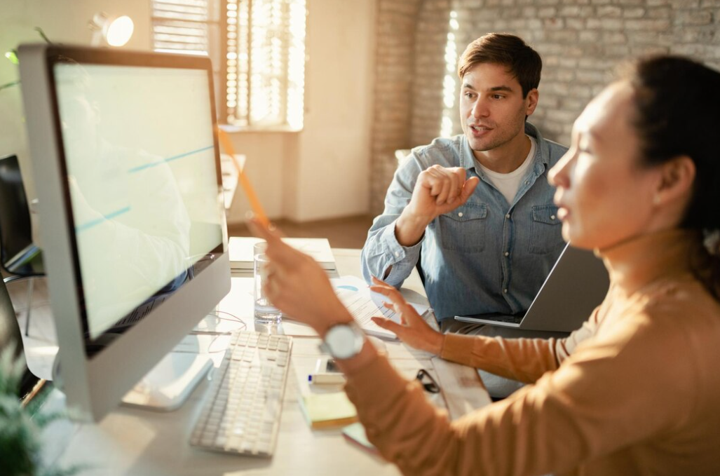 woman with watch sits in front of computer screen, man sits near her and looks on the screen